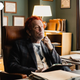 Malkovich sitting at a desk, wearing a jacket, vest, and t-shirt with a tie printed on it. His hair is dyed bright red.